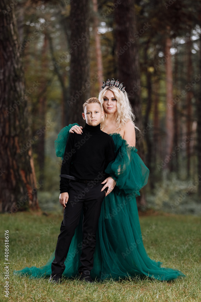 young beautiful blonde woman queen with young boy in black outfit. Princess mother walks with son. autumn green forest mystic. Vintage medieval shiny crown. Long evening green dress. magic fantasy