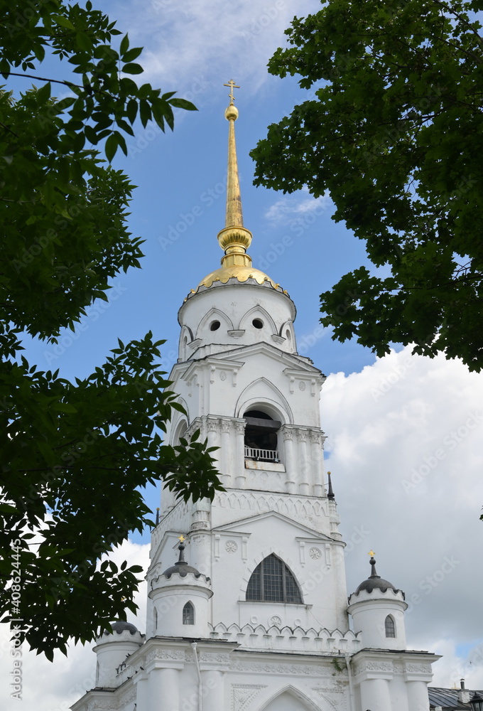 High white magnificent bell-tower with sharp golden spire and orthodox religious cross on top, belonging to old cathedral in Vladimir city, Russian 