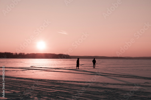 Horizontal landscape romantic outdoor photo with a silhouettes of a couple in love, walking through a snow-covered field during sunset against creamy red sky 