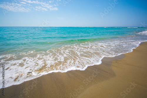 turquoise azure sea with a white wave rolls over a sandy beach on a clear sunny day against a blue sky