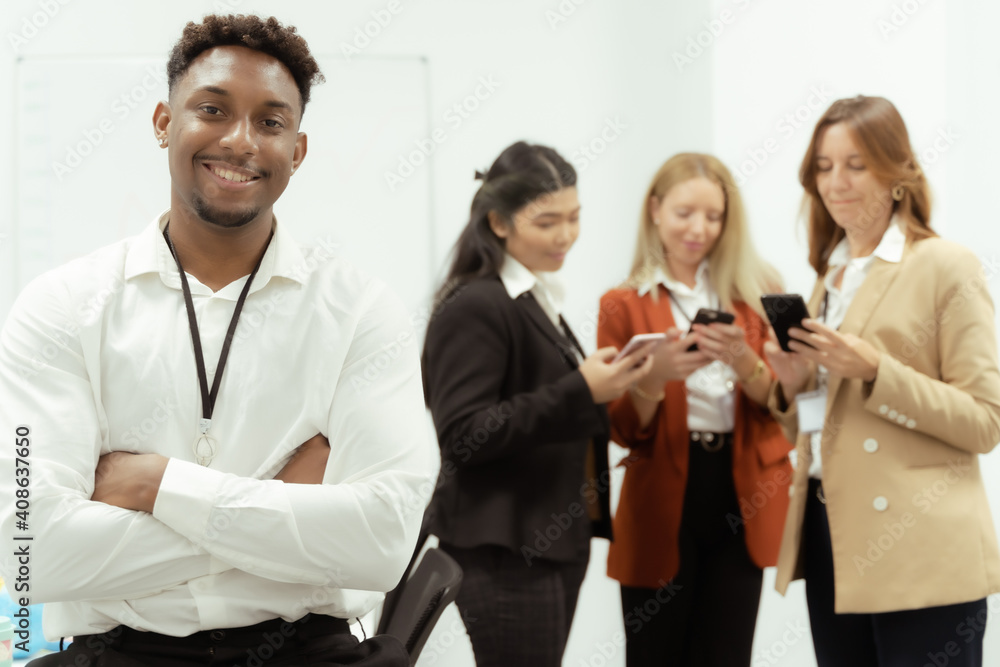 Young African male employee in formal clothes standing in the office. Group of people working behind with smartphones. Job concept.