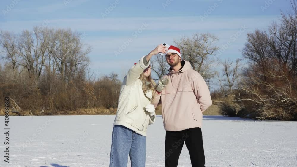 Young couple in christmas santa hats rides ice skating and takes a selfie on phone on frozen lake in forest Young couple in christmas santa hats rides ice skating and takes a selfie on phone on frozen lake in forest
