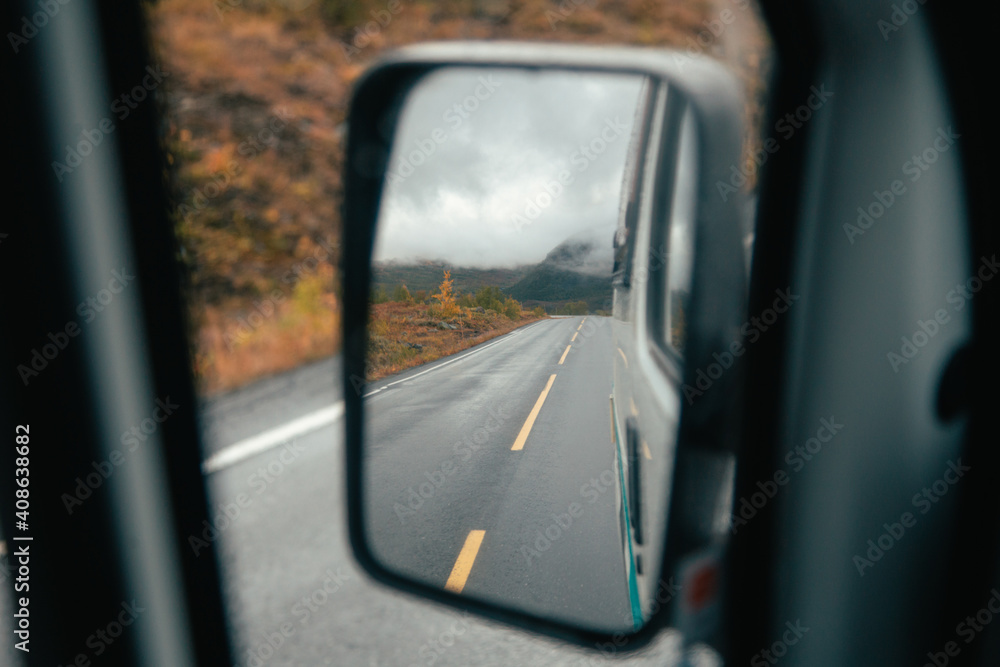 View from side mirror of camper van or rental car. Reflection of beautiful mountain road in autumn foliage during road trip to camping ground. Winter travel wanderlust concept