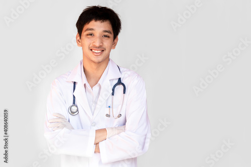A smiling doctor posing with arms crossed on a white background is wearing a stethoscope. Young Asian doctor wearing a white coat.