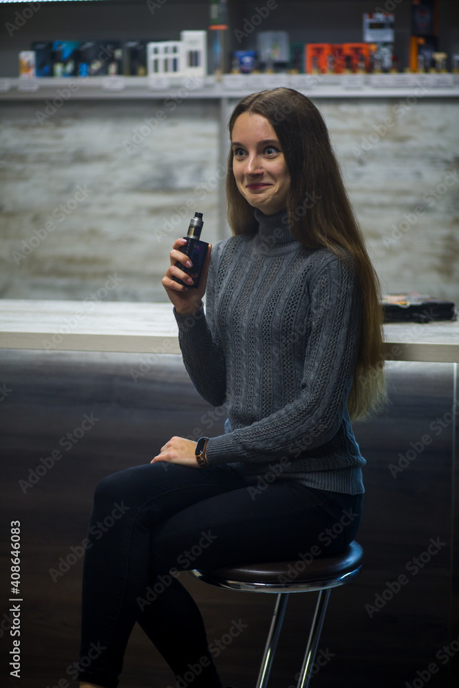Fototapeta premium Vaping teenager. Young pretty white caucasian teenage girl with long hair smoking an electronic cigarette indoors. Deadly bad habit. Vape activity.