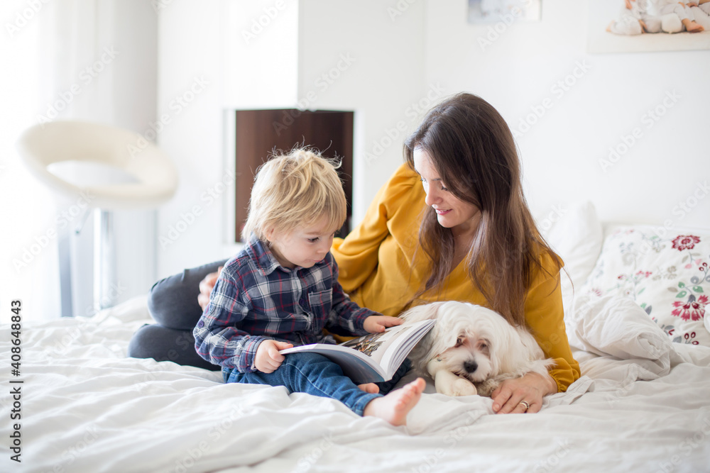 Mother, toddler child and puppy dog, lying on the bed, reading book together, family time Mother, toddler child and puppy dog, lying on the bed, reading book together, family time
