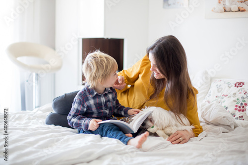 Mother, toddler child and puppy dog, lying on the bed, reading book together, family time