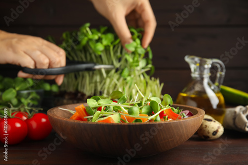 Woman cutting fresh organic microgreen at wooden table, focus on bowl of delicious salad