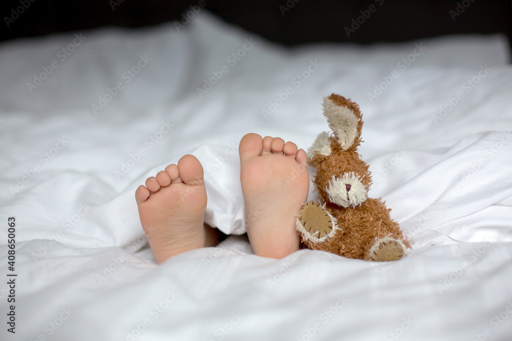 Little toddler child feet on the foreground, cute child, lying in bed, playing with little toy