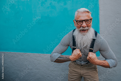 an older man in hipster clothes and glasses and a long white beard poses on a blue wall
