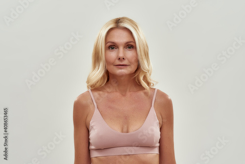 Human age. Portrait of a beautiful middle aged blonde woman in pink bra looking at camera while posing isolated over grey background