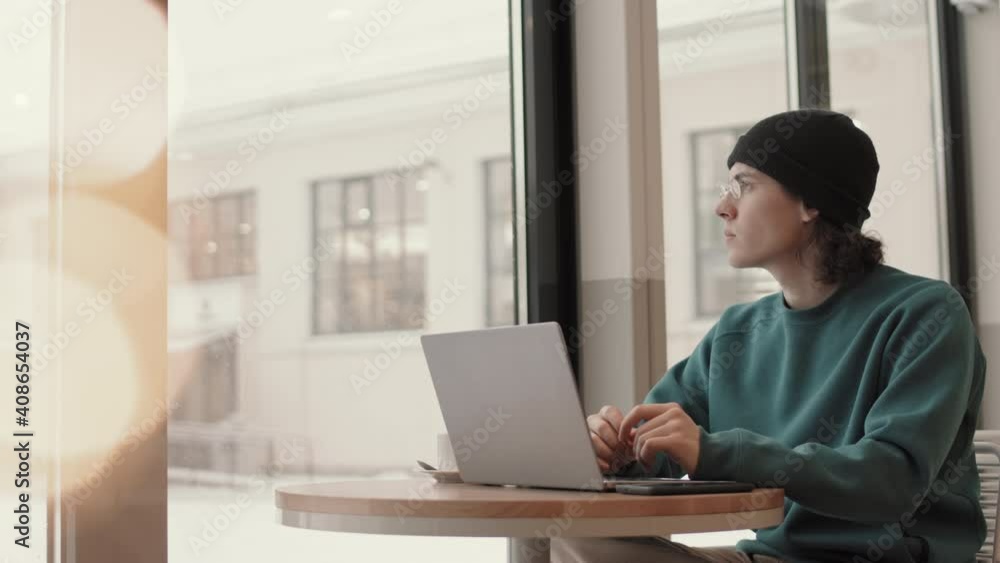 Lockdown of young hipster man wearing street wear and eyeglasses sitting at cafe table and working on computer Lockdown of young hipster man wearing street wear and eyeglasses sitting at cafe table and working on computer