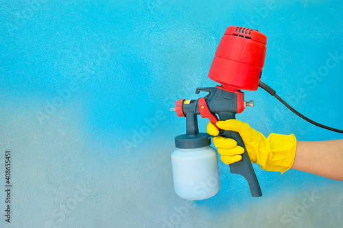 A worker's hand in a yellow glove holds a hand-held spray gun with an overhead compressor. Painting the wall of the house in blue. Household renovation. Copy space. Selective focus photo