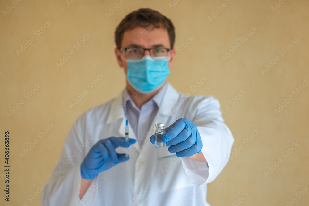 A man doctor in a protective mask and blue latex gloves holds an ampoule with a vaccine and a syringe. Focus on the ampoule A man doctor in a protective mask and blue latex gloves holds an ampoule with a vaccine and a syringe. Focus on the ampoule