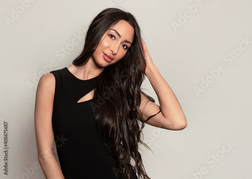 Beautiful young woman with long shiny black hair and natural make-up posing in dress on grey background 