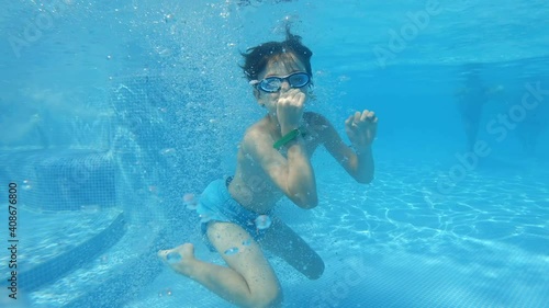 Happy boy jumping into a pool splashing in all directions and swimming under blue water smiling slow motion in a summer water park on a sunny day on vacation with his family. Travel. Tourism