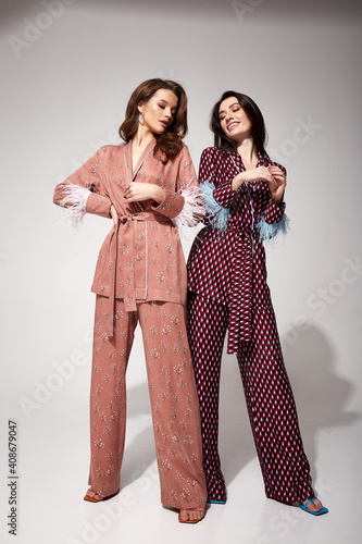 Two pretty sexy woman beautiful face natural makeup brunette hair wear silk textile suit pants and blouse with feathers comfort casual home dress luxury lifestyle pajama party glamour fashion model Fototapet