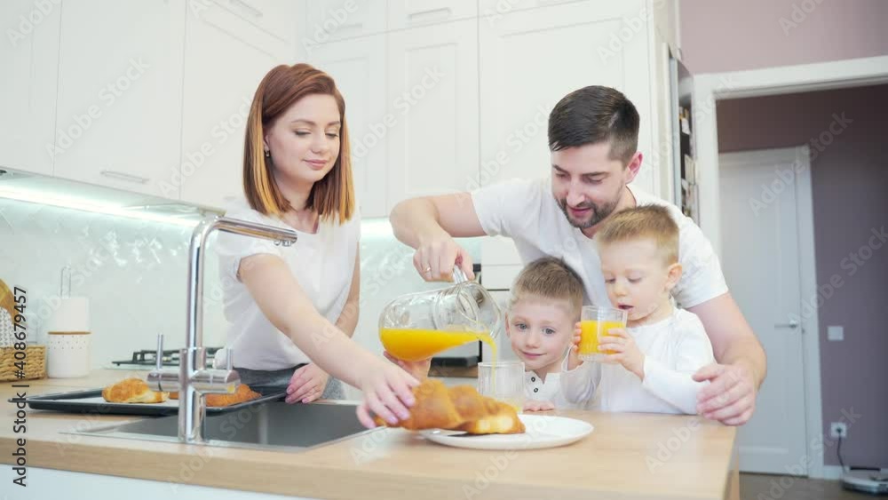 Young happy family in the kitchen preparing breakfast with young children. Mom and dad with little boys spend time together at home in the morning. Father pours the juice, hugs and kisses the mother Young happy family in the kitchen preparing breakfast with young children. Mom and dad with little boys spend time together at home in the morning. Father pours the juice, hugs and kisses the mother