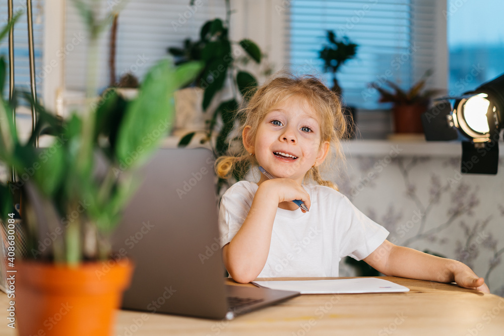 Happy little girl vlogger sitting at laptop looking at camera, smiling and making online video call video blog recording, portrait Happy little girl vlogger sitting at laptop looking at camera, smiling and making online video call video blog recording, portrait