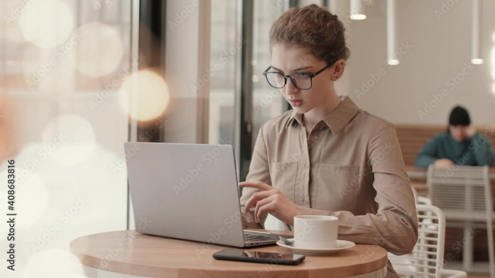 Lockdown of young attractive Caucasian businesswoman wearing beige shirt and eyeglasses sitting at cafe table and working on laptop Lockdown of young attractive Caucasian businesswoman wearing beige shirt and eyeglasses sitting at cafe table and working on laptop