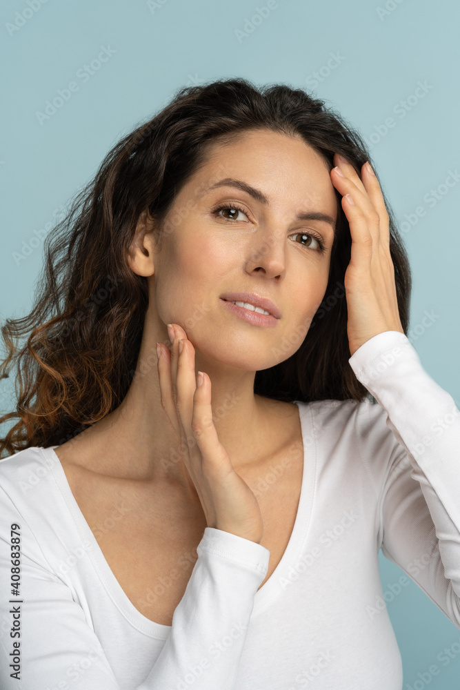Studio portrait of young woman with natural makeup, curly hair, touching well-groomed pure skin on face, isolated on blue background. Beauty, facelift, anti-aging cosmetic procedure. Studio portrait of young woman with natural makeup, curly hair, touching well-groomed pure skin on face, isolated on blue background. Beauty, facelift, anti-aging cosmetic procedure.