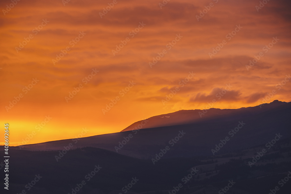 Atmospheric landscape with dark silhouettes of mountains on background of vivid orange dawn sky. Colorful scenery with sunset or sunrise of illuminating color. Sundown paysage with sunbeam on mountain