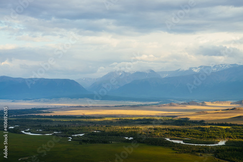 Scenic landscape with vast plateau with mountain river and forest in sunlight on background of snowy mountain ridge under cloudy sky. Green mountain valley in sunshine and mountain range on horizon.
