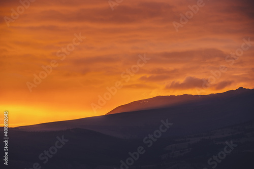 Atmospheric landscape with dark silhouettes of mountains on background of vivid orange dawn sky. Colorful scenery with sunset or sunrise of illuminating color. Sundown paysage with sunbeam on mountain © Daniil