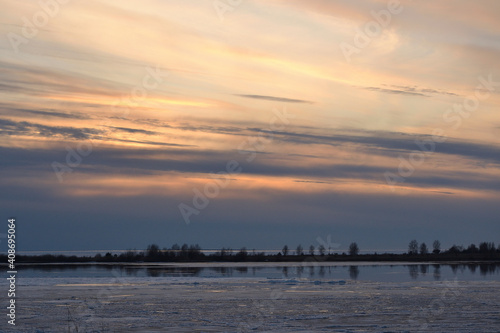 A landscape view of the frozen sea during ice drift in spring with dramatic sky and with reflections in melt water in the evening at sunset. In the distance, the black strip of the island. © Helena
