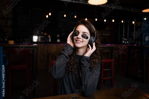 a girl sits at a table in a cafe with black patches on her face and headphones on her head, listening to music with great difficulty, smiling with pleasure