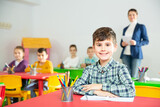 Cute intelligent schoolboy sitting in classroom on background with classmates and teacher