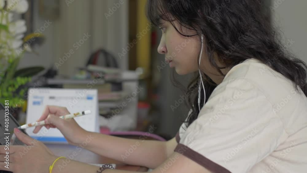Close up asian female teenager writing on digital tablet at the desk for lesson online at home. University girl in casual dress with earphone doing homework on the table. Study of the exam. Close up asian female teenager writing on digital tablet at the desk for lesson online at home. University girl in casual dress with earphone doing homework on the table. Study of the exam.