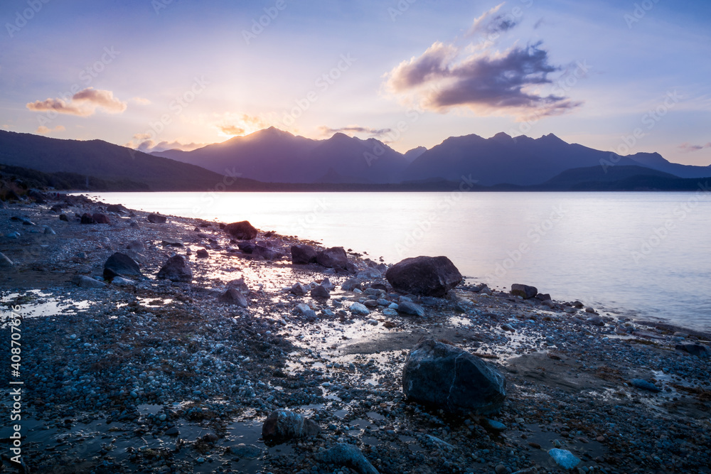 Colorful sunlight behind the mountain with reflections in the water and water pools on the shore at Lake Te Anau - Patience Bay - in Fiordland National Park, New Zealand, South Island. Colorful sunlight behind the mountain with reflections in the water and water pools on the shore at Lake Te Anau - Patience Bay - in Fiordland National Park, New Zealand, South Island.