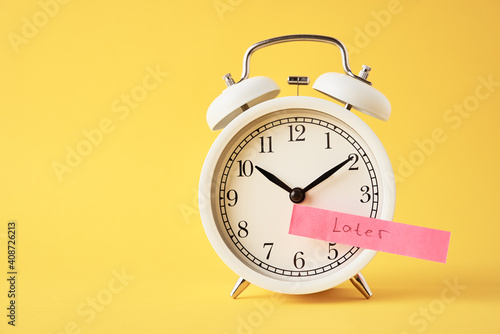 Procrastination and delay concept. Sticky note with word later on white alarm clock on yellow background. Urgency time