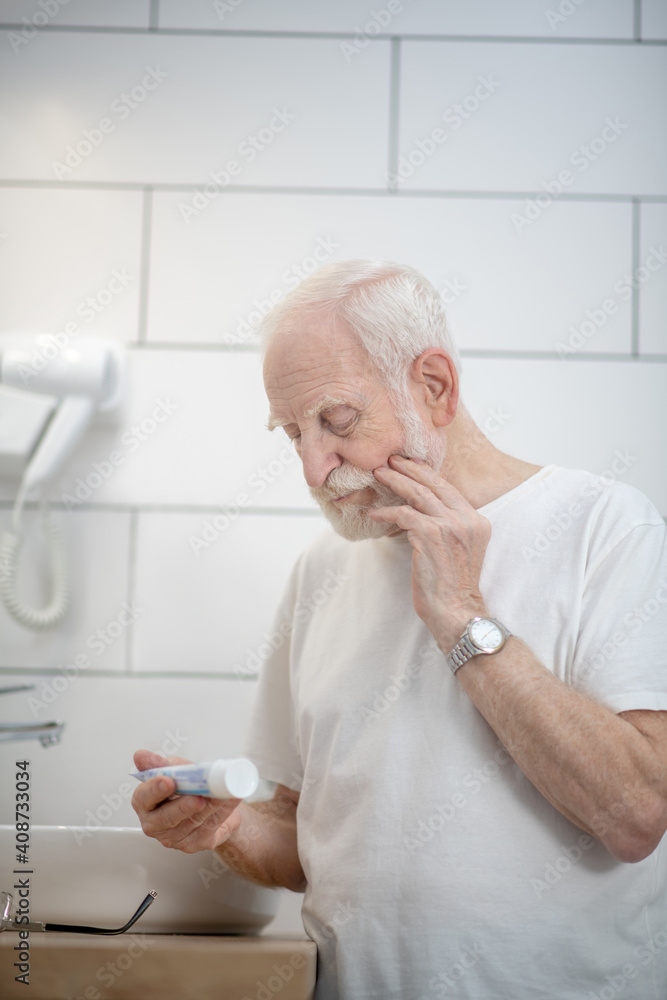 Gray-haired man in white tshirt holding a cream and looking thoughtful Gray-haired man in white tshirt holding a cream and looking thoughtful