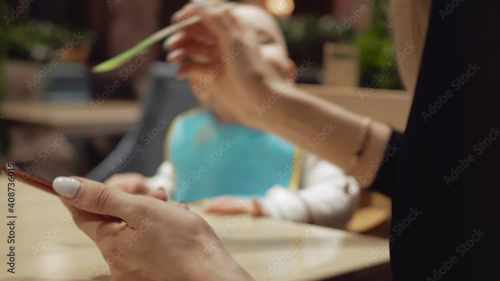 A woman with blond hair, dressed in a black blouse, holding a smartphone in her left hand, feeding a child in a blue bib, the child willingly eats, opening her mouth and looking around A woman with blond hair, dressed in a black blouse, holding a smartphone in her left hand, feeding a child in a blue bib, the child willingly eats, opening her mouth and looking around
