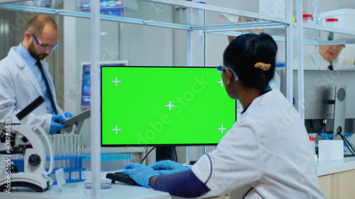 African doctor working at computer with green screen in modern equipped lab. Multiethnic team of microbiologists doing vaccine research writing on device with chroma key  isolated  mockup display.