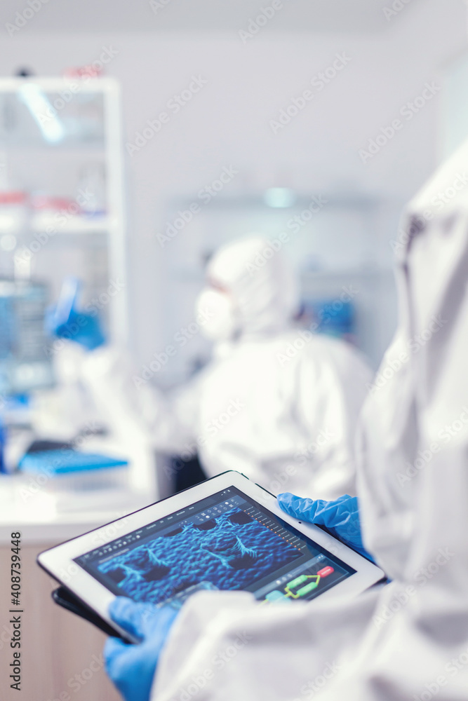 Chemist dressed in protection suit for coronavirus holding tablet pc in laboratory. Team of scientists conducting vaccine development using high tech technology for researching treatment against Chemist dressed in protection suit for coronavirus holding tablet pc in laboratory. Team of scientists conducting vaccine development using high tech technology for researching treatment against