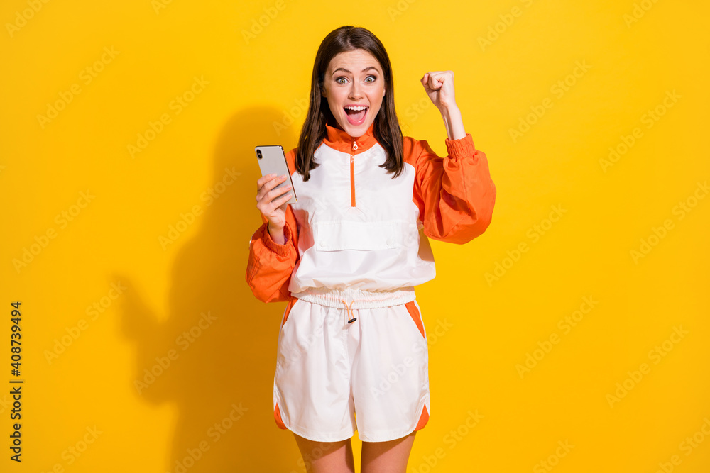 Photo portrait of celebrating woman with raised fist holding phone in one hand isolated on vivid yellow colored background Photo portrait of celebrating woman with raised fist holding phone in one hand isolated on vivid yellow colored background