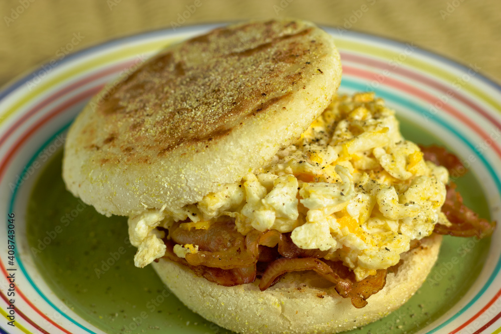 Toasted english muffin with fried bacon and scrambled eggs on a colorful plate.