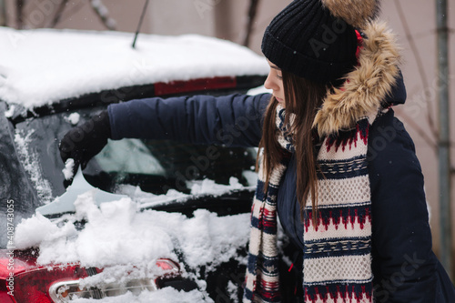 A woman is cleaning snowy window on a car with snow scraper. Pretty woman warmy dressed clean her car outdoors. Cold snowy and frosty morning. Black car photo