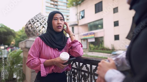 Asian muslim woman enjoy talking with friend, Islam community, holding coffee cup, taking a break at city downtown, outdoor coffee shop, traffic behind showing urban street background, smiling and fun photo