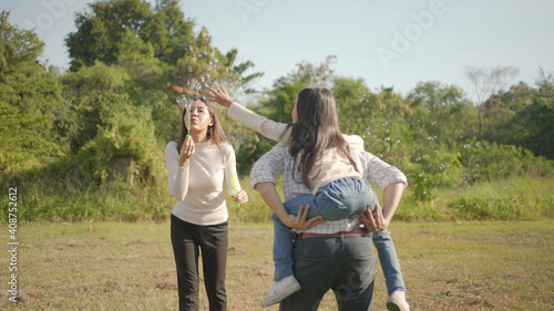 Happy young asian father piggyback with his little girl while mother run to play with her in the park and enjoyed ourselves together in the green garden. Family enjoying sunny fall day in nature.