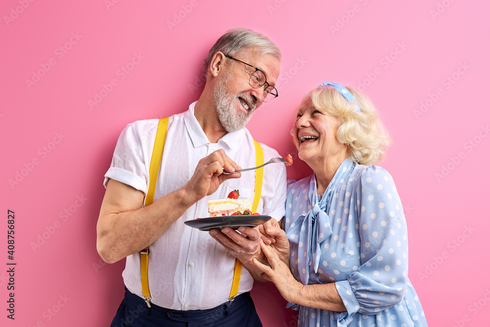 senior man treats a woman with a cake, elderly couple celebrating birthday or anniversary, isolated on pink background, portrait