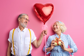senior couple celebrating Valentine's Day isolated on pink background. beautiful woman and man in party clothes, holding gift box and air balloon in heart shape