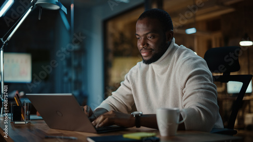 Handsome Black African American Man Having an Online Conversation on a Laptop Computer in Creative Office Environment. Happy Male is Browsing Social Media and Replying to Friends in Messenger.