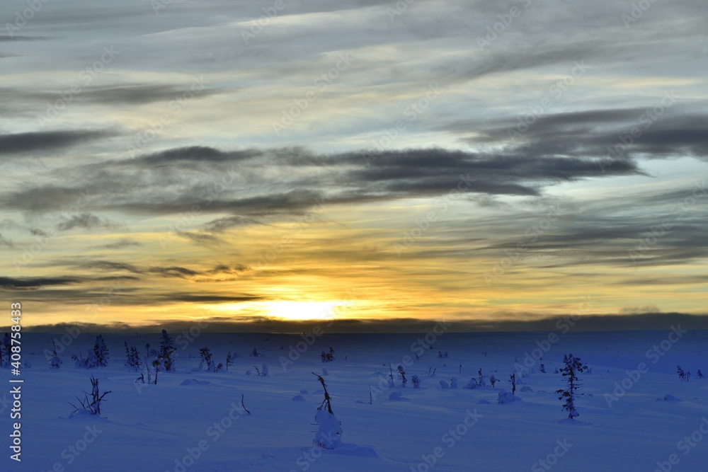 A winter landscape with snow and trees in a colorful sunset with dark clouds in the sky.