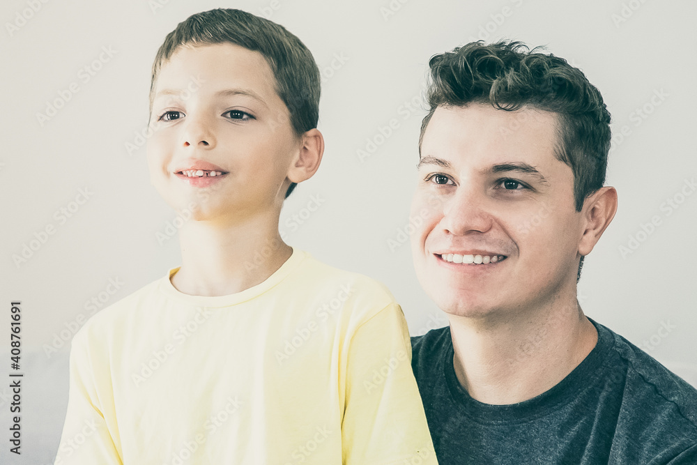 Happy boy and his dad sitting together indoors and looking away. Medium shot. Family and parenthood concept Happy boy and his dad sitting together indoors and looking away. Medium shot. Family and parenthood concept