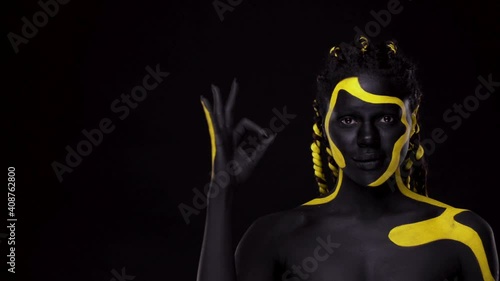 Face art. Girl showing OK sign with fingers. Woman with black and yellow body paint. Young african girl with colorful bodypaint. An amazing afro american model with makeup. photo