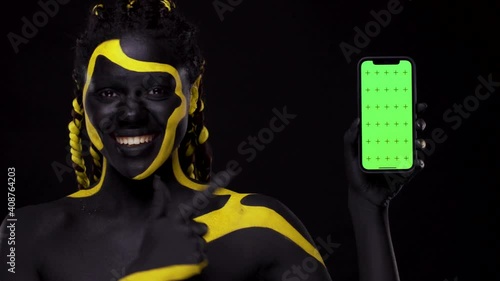 Face art. Woman with black and yellow body paint. Girl pointing finger at mobile phone screen with chroma key. Young african female with colorful bodypaint. An amazing afro american model with makeup. photo